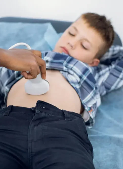 young boy lays on an exam table with plaid shirt pulled over his abdomen, while ultrasound tech uses handheld device to view ultrasound of child's abdomen