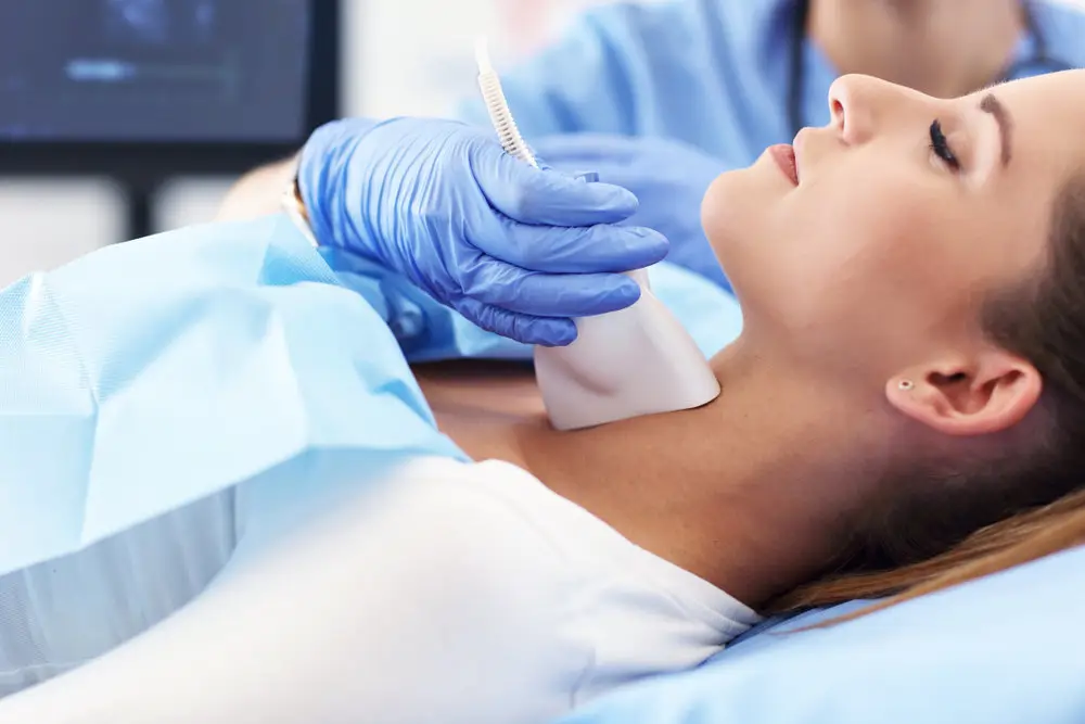 ultrasound tech wearing gloves uses a handheld device to view ultrasound of woman's thyroid gland
